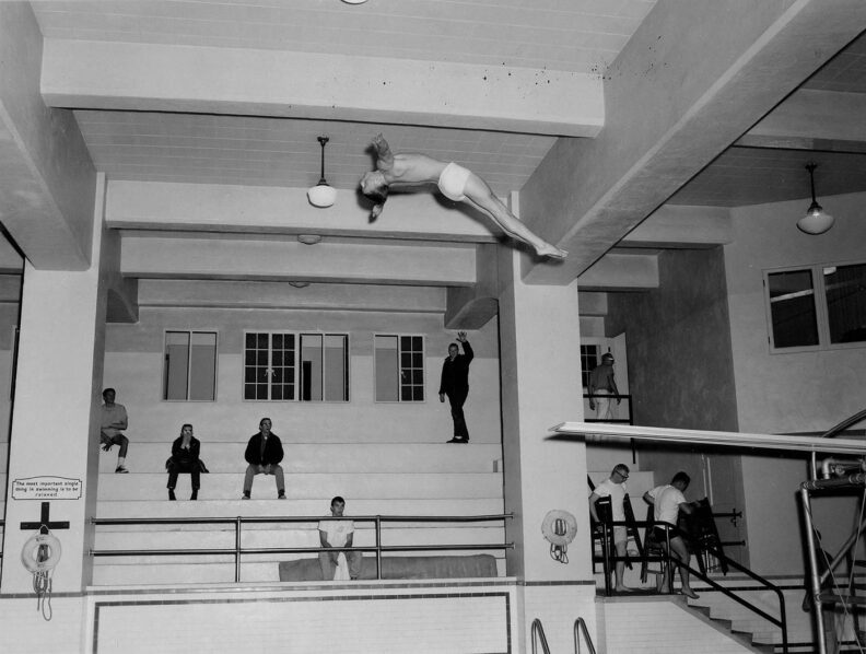Black and white photo of a young man in swim trunks diving into a pool during a competition