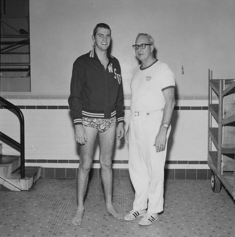 black and white photo of a smiling young man in swim trunks and WSU jacket next to a middle-aged man with horn rim glasses, white t-shirt, and white pants
