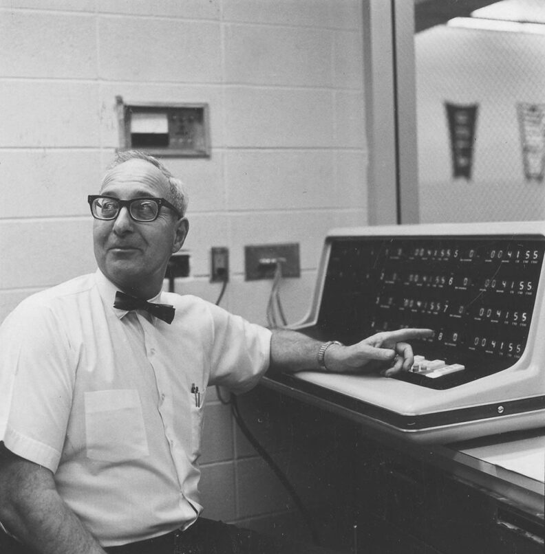 Middle-aged man in 1970 black and white photo, wearing horn rim glasses and bowtie, points to an electronic timekeeping machine.