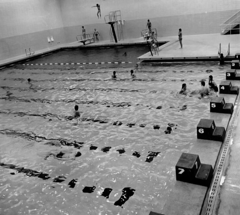 Black and white photo of swimmers in a pool with racing lanes and diving boards