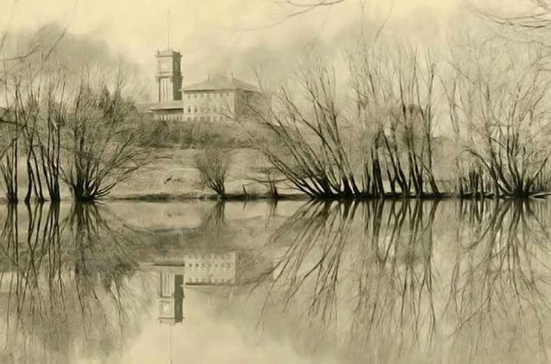 Sepia tone photo from the 1920s of a small lake lined with trees and brick college buildings and clock tower in the background.