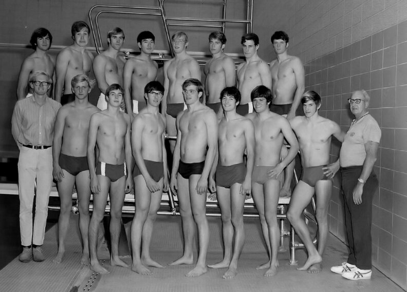 black and white photo of a group of young male swimmers in trunks, with two coaches