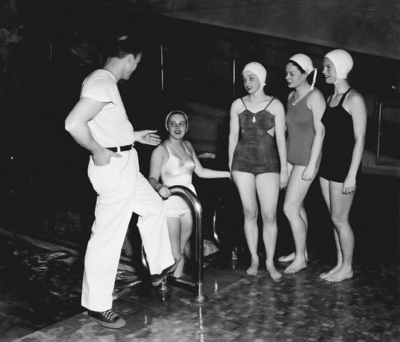Black and white photo of four young woman swimmers and an older male coach next to a pool