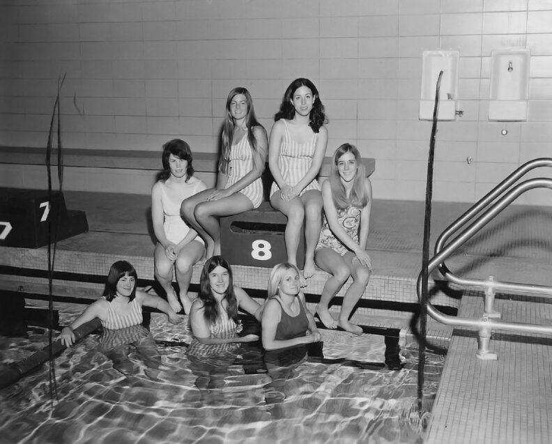 A group of seven young women swimmers sit in and next to a pool