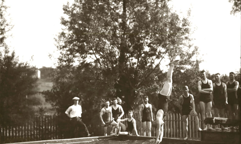 Black and white photo from 1917 of a young man diving into a pool with other young men watching from the side
