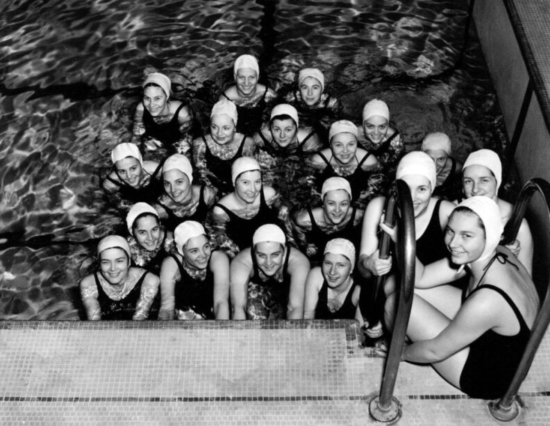 Black and white photo of young women in bathing suits and caps in 1937 gathered by the edge of the pool