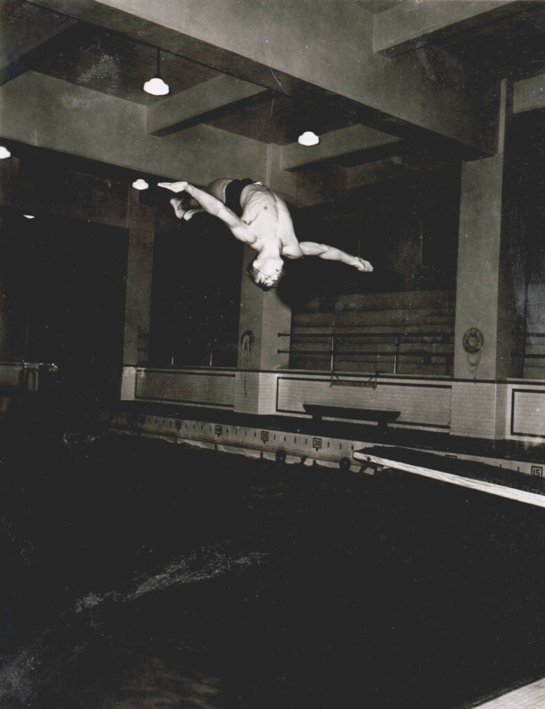 Black and white photo of a young man diving backwards into a pool.
