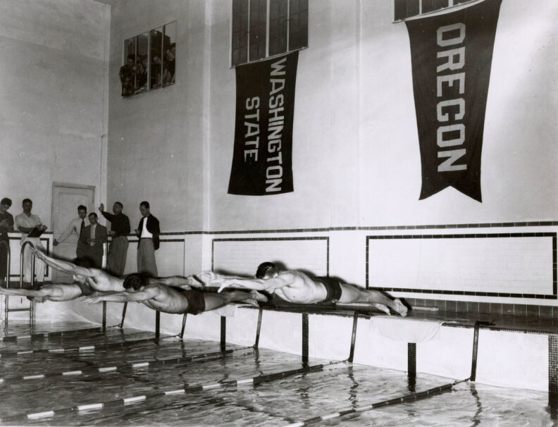 Black and white photo from 1952 of male swimmers jumping into a pool for a race. Banners on the wall say Washington State and Oregon.