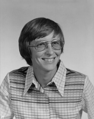 Black and white closeup of a smiling woman with short hair, glasses, and plaid shirt