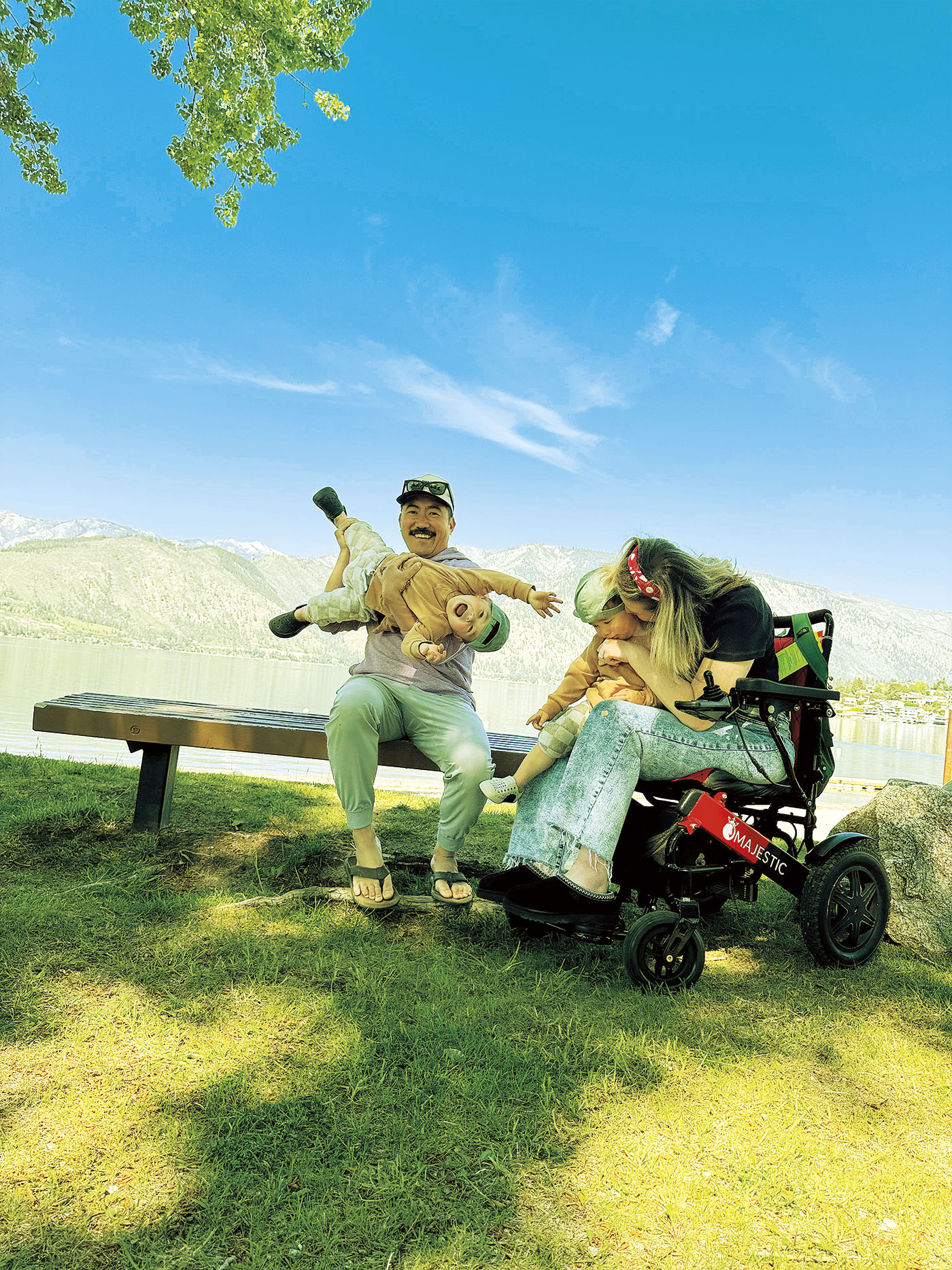 Man with baseball cap on a bench holds a laughing toddler and woman in wheelchair hugs a toddler