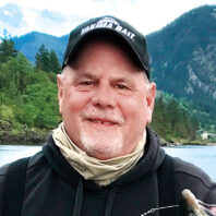 Headshot of man with gray goatee in baseball cap, next to a river and mountains