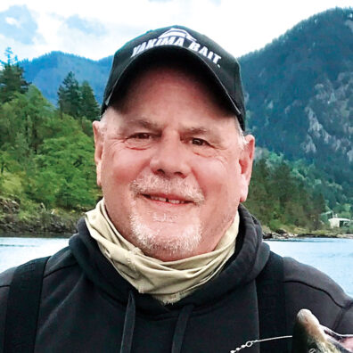 Headshot of man with gray goatee in baseball cap, next to a river and mountains