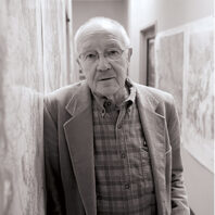 Bill Lipe as an older man in suit jacket and plaid shirt leans against a wall covered with maps