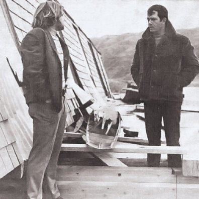 Two men in black and white photo in front of ruined dockhouse