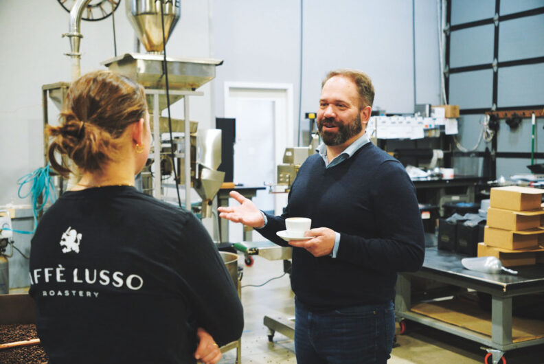 Man stands with a coffee cup on a saucer talking to employee at coffee roasting space in background