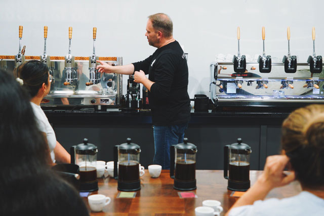 Man points to coffee grinding equipment during a coffee tasting event. People in foreground at table have french press coffee makers