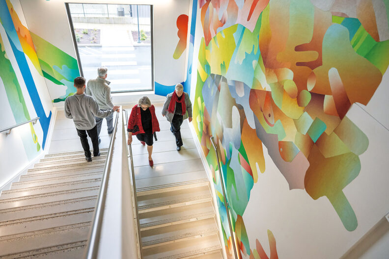 WSU President Elizabeth Cantwell goes down some stairs at WSU Vancouver with three other people. A colorful mural is on the wall.