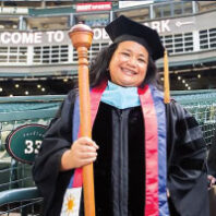 Rosie Rimando-Chareunsap in graduation gown as college president, holding a gonfalon