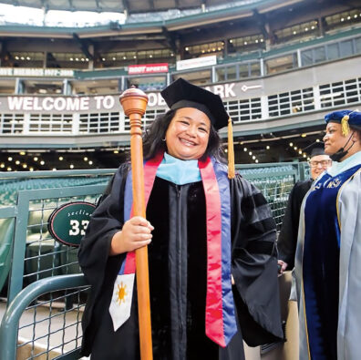 Rosie Rimando-Chareunsap in graduation gown as college president, holding a gonfalon