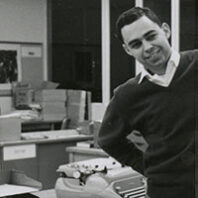 Black and white photo of a young college-age smiling man in a newspaper editorial room