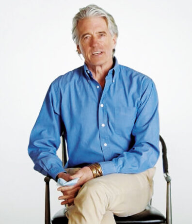 Man with gray hair in blue dress shirt, holding a paper mask, sits in a chair as he talks.