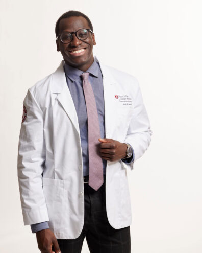 Joel Bervell smiling, wearing white doctor's coat, tie and dress shirt