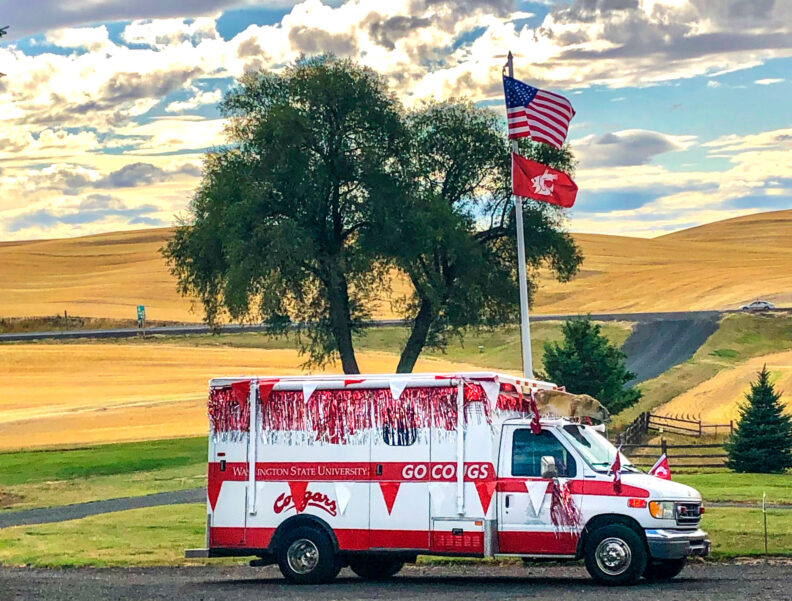 Converted ambulance decorated in Washington State University logos and colors in front of a grain field
