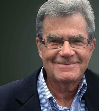 Close-up color photograph of an older adult wearing glasses, a striped collared shirt, and a dark jacket, facing the camera against a dark green background.