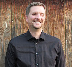 Portrait of a bearded man wearing a black button-up shirt, standing against a weathered wooden wall
