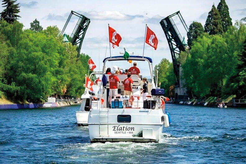 Boat traveling through a waterway with people onboard and drawbridges raised in the background, flying WSU flags