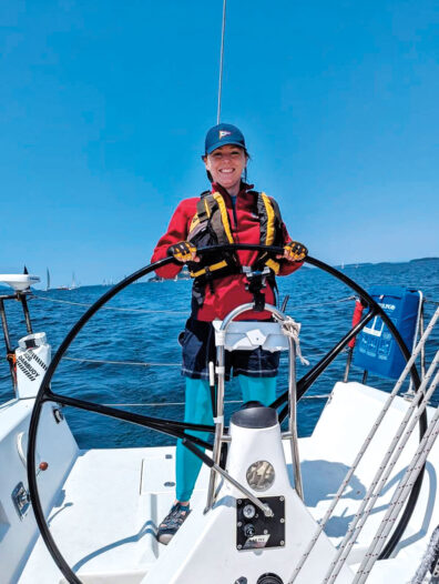 Woman standing at the wheel of a sailboat on open water under a clear blue sky.