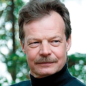 Close-up outdoor portrait of an older man with short hair and a mustache, wearing a dark high-collared shirt, with blurred green foliage in the background.