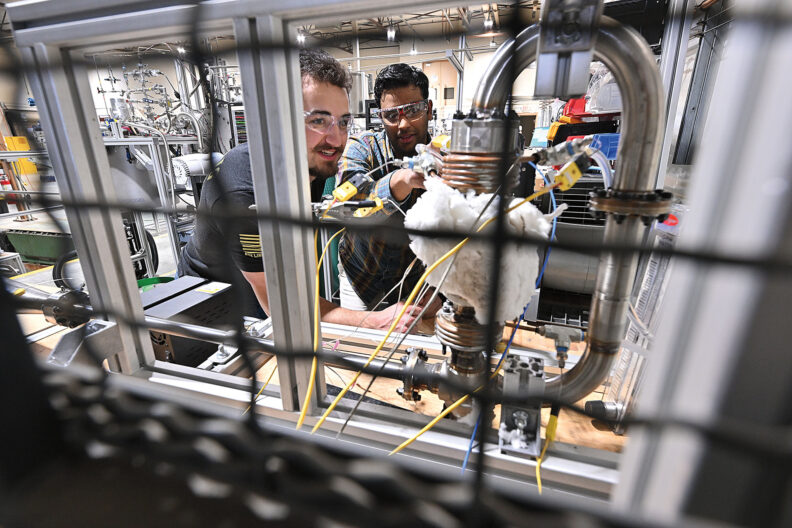 Two engineers working inside an industrial laboratory setup, examining pipes, sensors, and wiring within a metal-framed testing structure.