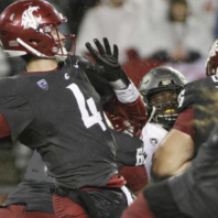 Action photograph of a quarterback in a dark uniform and helmet throwing a football during a game, with opposing players and teammates nearby in rainy conditions.