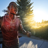 Bronze statue of a runner wearing a WSU singlet, mid‑stride on a campus walkway, lit by low sunlight with trees and buildings in the background.