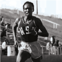 Black‑and‑white photo of a runner wearing a WSU uniform, mid‑stride on a track with stadium seating in the background