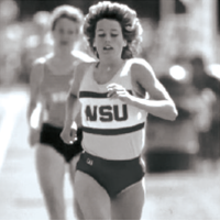 Black‑and‑white photo of a runner wearing a WSU uniform, mid‑stride on a track with stadium seating in the background.