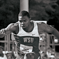 Black‑and‑white photo of a runner wearing a WSU uniform, mid‑stride on a track with stadium seating in the background.