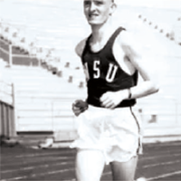 Black‑and‑white photo of a runner wearing a WSU uniform, mid‑stride on a track with stadium seating in the background.