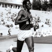 Black‑and‑white photo of a runner wearing a WSU uniform, mid‑stride on a track with stadium seating in the background.
