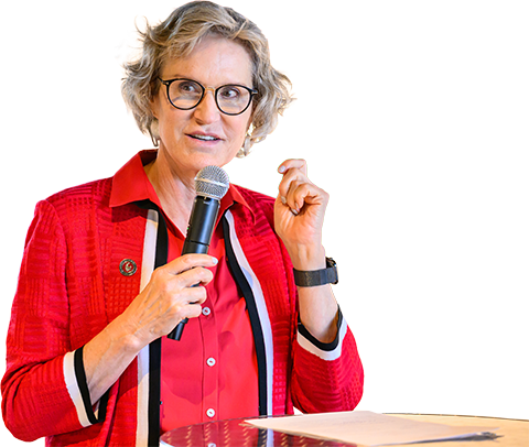 Woman speaking at a podium while holding a microphone, wearing a red jacket and glasses, against a plain white background.