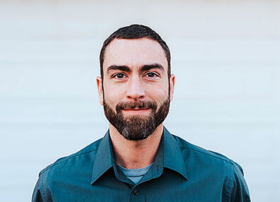Close-up portrait of a man with short dark hair and a beard, wearing a buttoned shirt, standing in front of a softly blurred light-colored background.