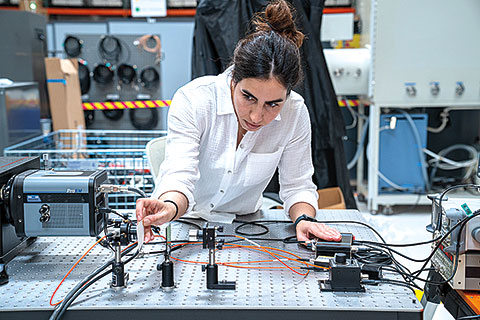 Woman researcher leaning over an optical table, adjusting cables and instruments in a laboratory filled with scientific equipment and control panels.