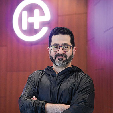 Portrait of a man with glasses and a beard standing with arms crossed, positioned in front of a glowing circular logo on a wall.