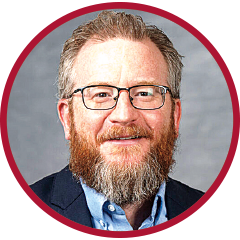 Circular headshot of a man with glasses and a full beard, smiling slightly, framed by a red border against a neutral background.