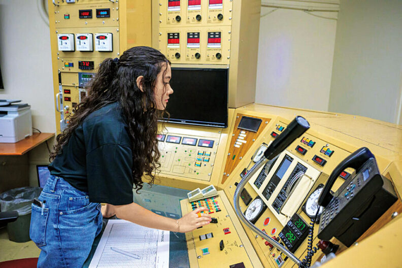 Woman operator standing at a large control console, pressing buttons and viewing analog gauges and screens in a technical control room.