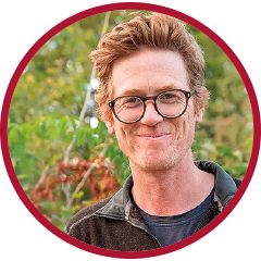 Circular outdoor headshot of a man wearing a zip-front jacket, framed by a red border with foliage in the background.