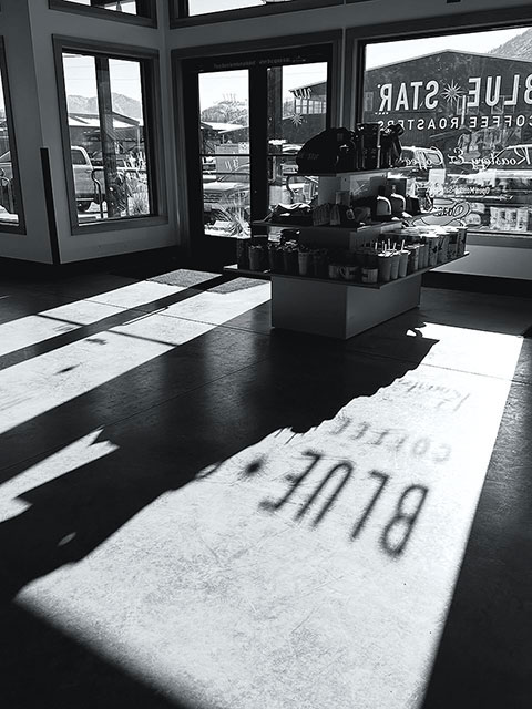 Sunlit interior of a coffee shop with large windows, a central merchandise table, and shadows cast across the floor.