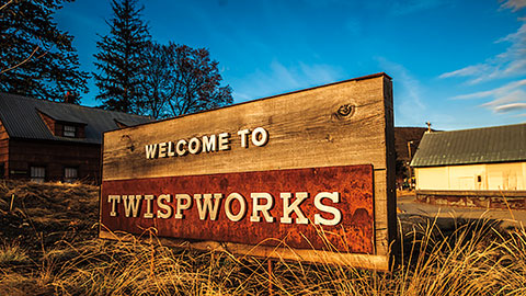 Wooden roadside sign reading ‘Welcome to TwispWorks,’ set among grass and rustic buildings under a blue sky.