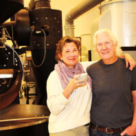 Woman and man standing together inside a coffee roasting facility, with large industrial roasting equipment in the background.